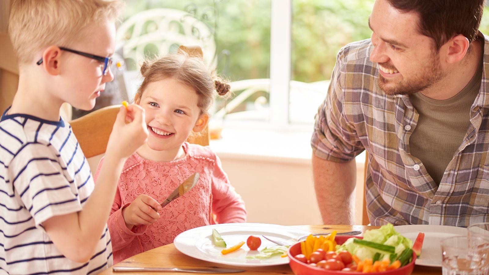 A father with young son and daughter watching the son try a piece of a vegetable.